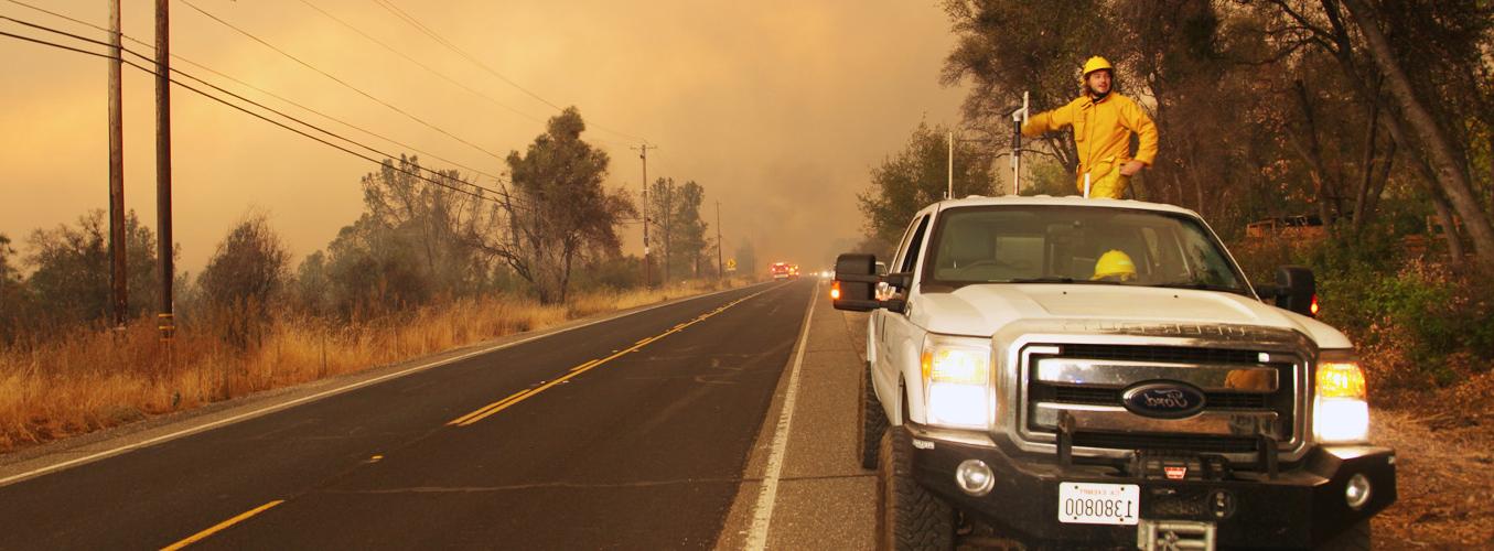 Firefighter in white truck on side of road with fire and smoke in background.